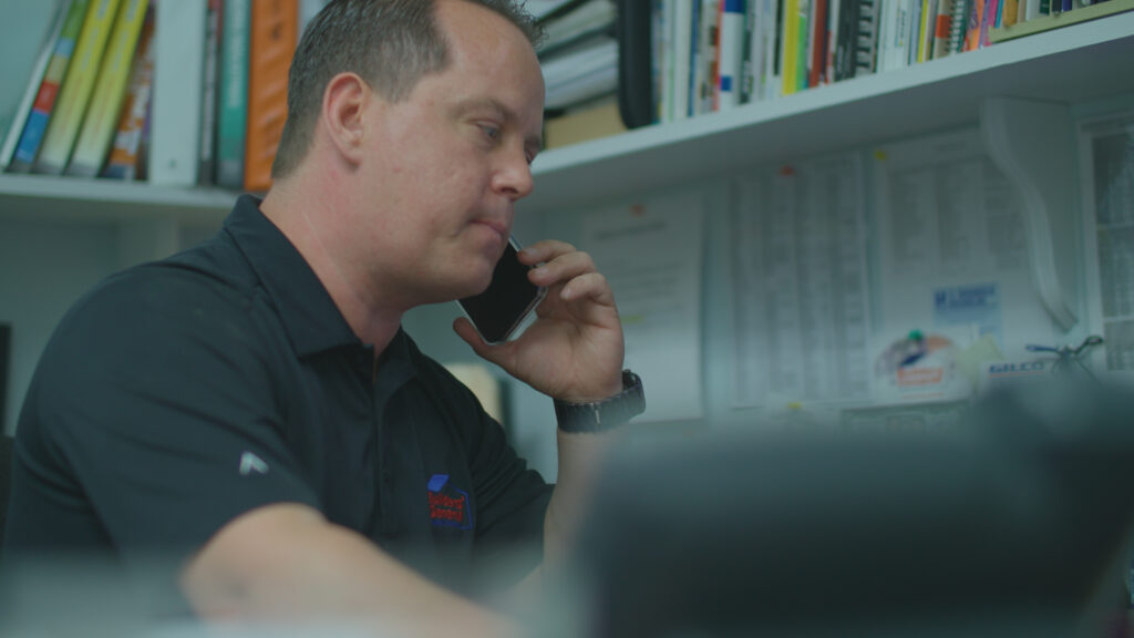 Builders’ General`s Operations Manager Dan Whitmeyer sitting in his office at Builders’ General Little Silver location. He has his phone against his left ear and there are books on a shelf in the background.