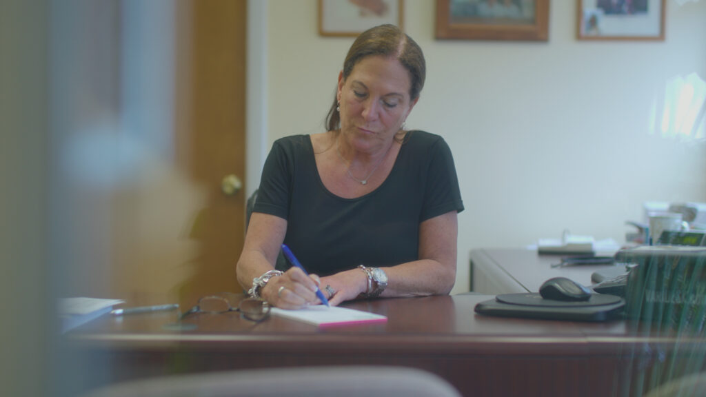 Builder’ General Vice President Betsy Shaheen sitting at her desk. She is writing on a notepad. Her glasses are next to the notepad. 