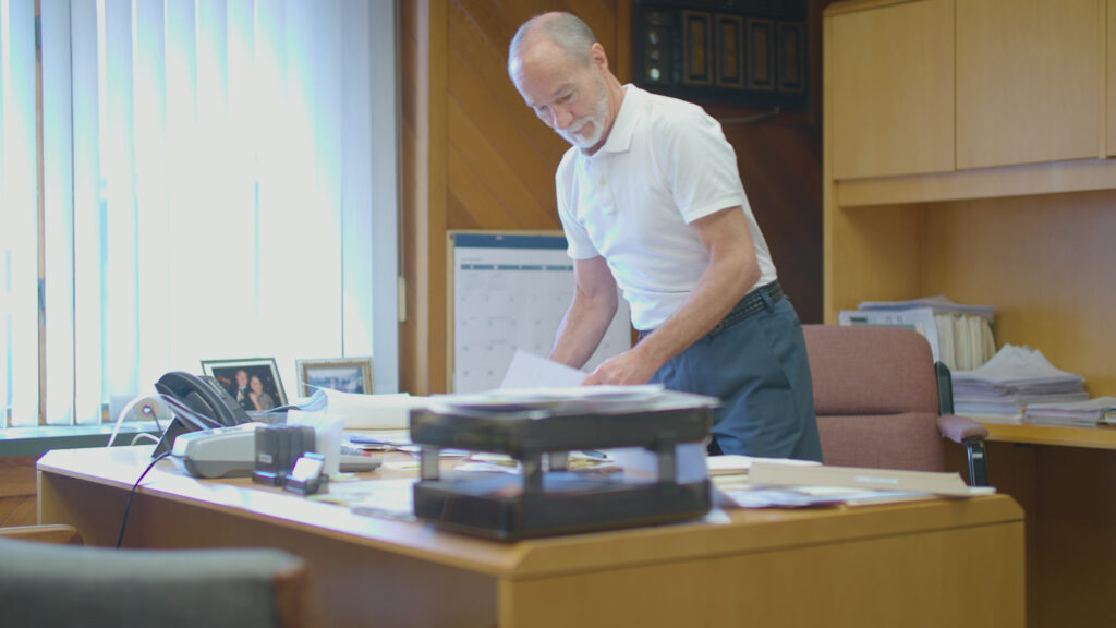 Builders’ General`s employee standing over his desk at Builders’ General Little Silver location. Dressed in white polo and dress slacks, he is shuffling papers around a cluttered light wooden desk. 