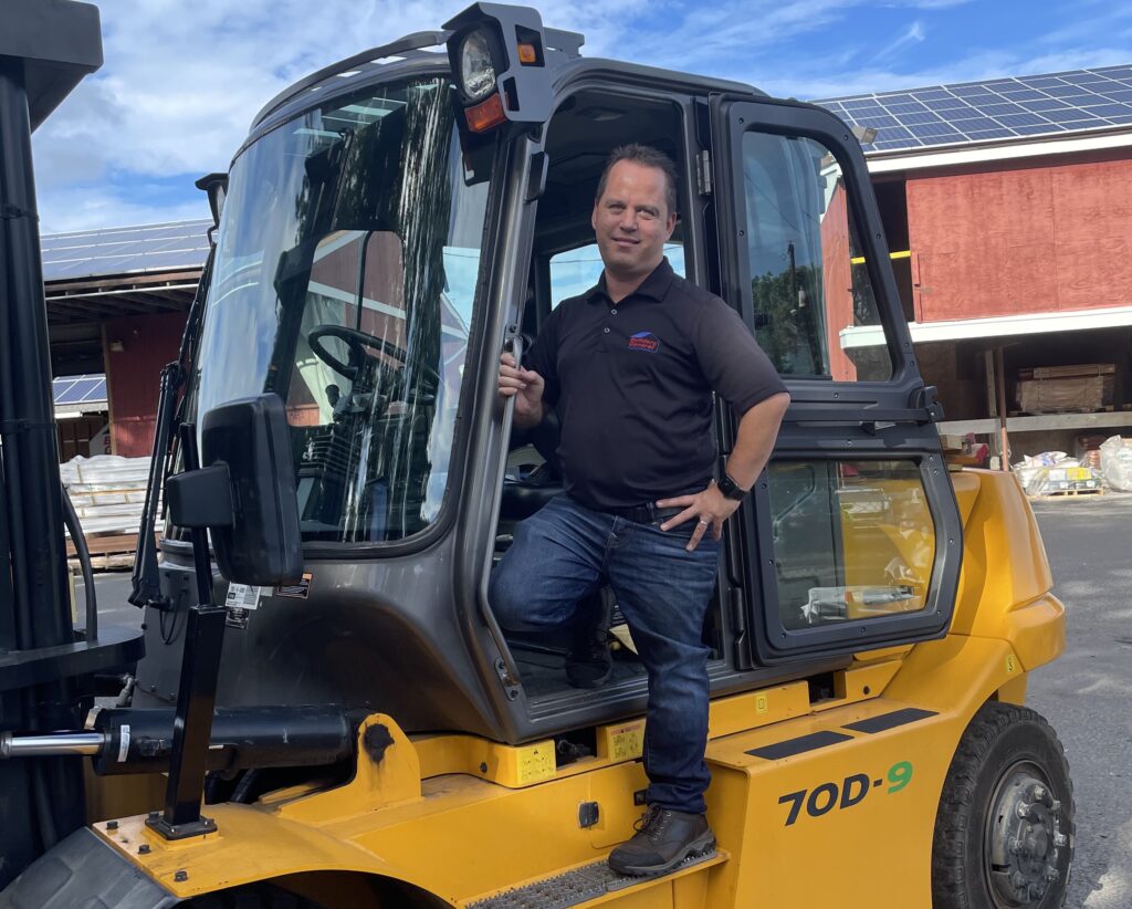 Dan Whitmeyer is smiling while positioned on the step of a forklift outside the Builders’ General Little Silver lumberyard. Lumber and building materials are stacked in the background.