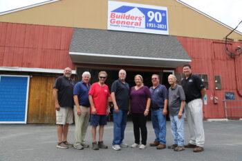 8 Builders’ General employees stand in front of the red and yellow building with a company sign above them saying “90th” for their 90th anniversary.